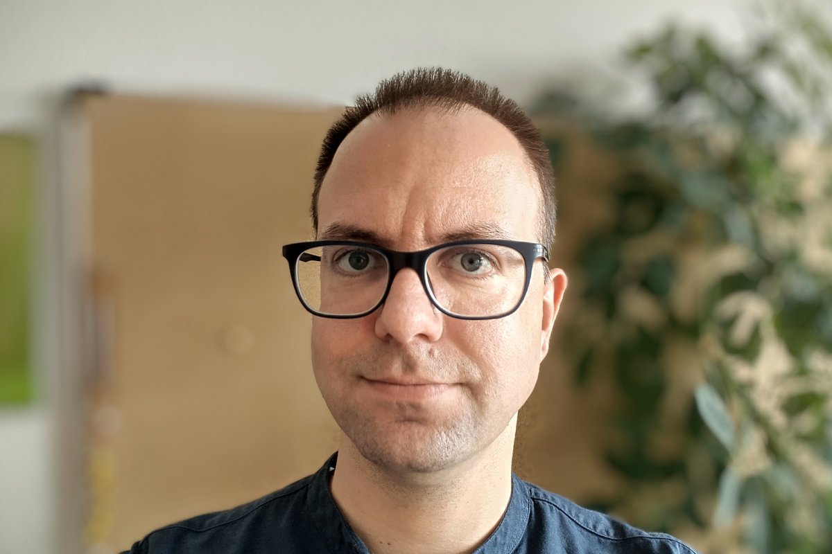 Porträt eines Mannes mit Brille und dunklem Hemd vor unscharfem Hintergrund mit Pflanze